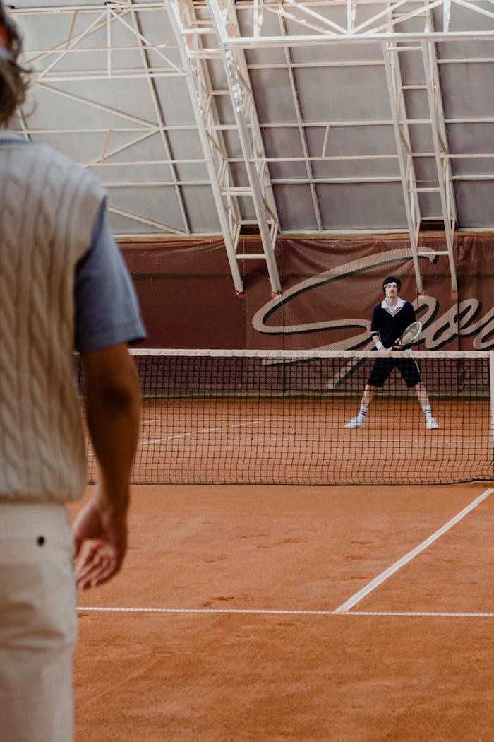 about-img-02 Two men playing tennis indoors on a clay court with stylish vintage attire.