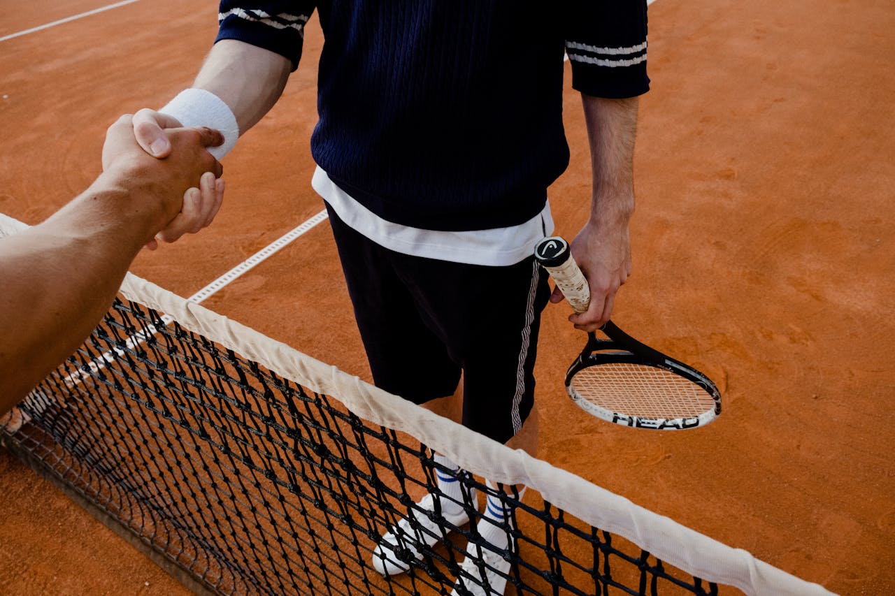 gallery-01 A close-up of tennis players shaking hands over the net on a clay court.