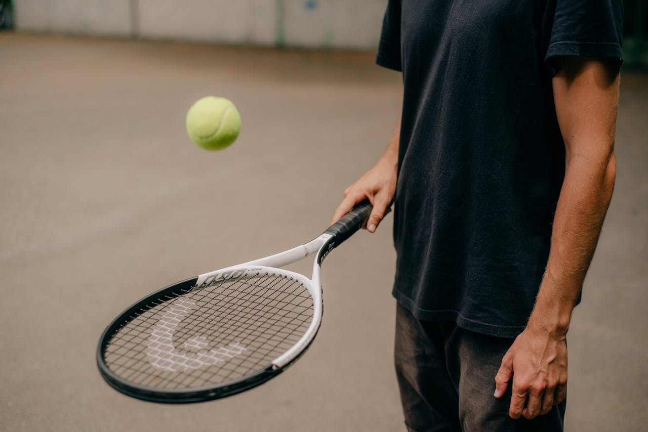 gallery-03 A man in a black t-shirt skillfully handling a tennis racket with a ball in motion outdoors.